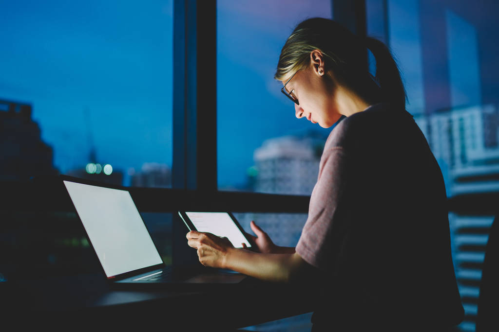 Молодая женщина-фрилансер в очках смотрит тренировочный вебинар на веб-странице на сенсорной панели, подключенной к беспроводному интернету в темноте дома. Женщина обновляет программное обеспечение на ноутбуке с чистым экраном
 - Фото, изображение