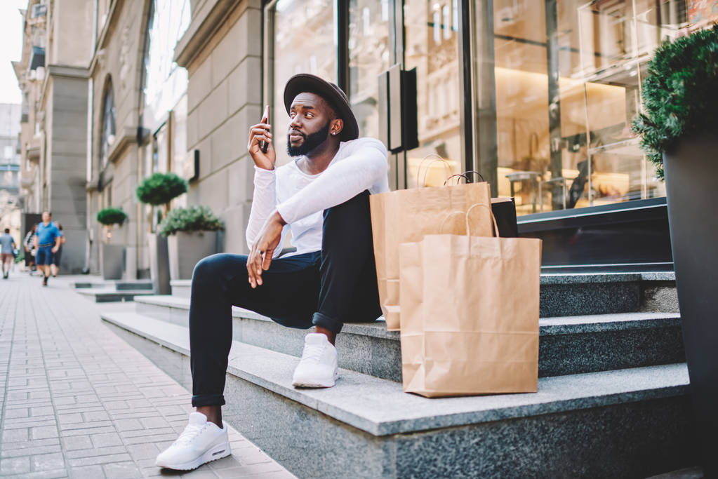 Grave afro-americano maschio shopper fare telefonata per contattare operatore di servizi in attesa di consegna, giovane uomo dalla pelle scura seduto sulle scale del negozio vicino a sacchetti di carta con spazio per l'etichetta
 - Foto, immagini