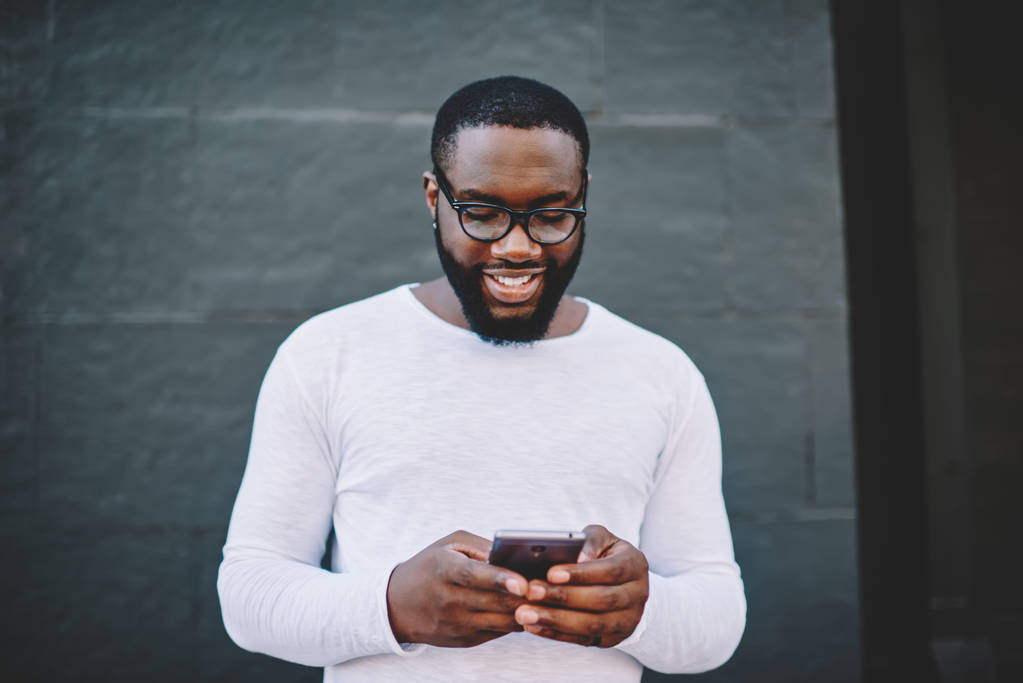 Cheerful african american man in spectacles searching cafes and restaurants in application on mobile,smiling dark-skinned guy reading news from networks on phone standing on copy space for advertising - Photo, Image