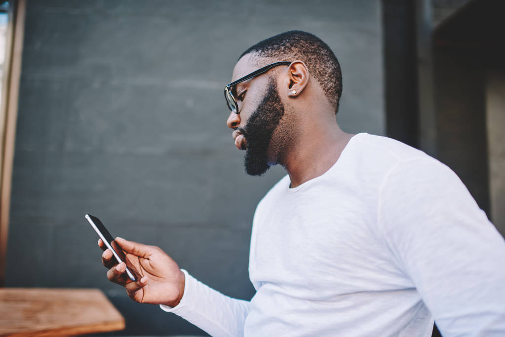 Boční pohled fešák afroamerické čtení zpráv na telefonu stoje venku v tmavou bokové v košili s kopií prostor pro obchodní název nebo označení instalaci aplikace na mobil - Fotografie, Obrázek