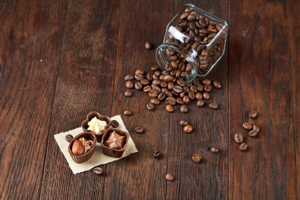 Μερική άποψη του αναποδογυρισμένο γυάλινο βάζο με κόκκους καφέ και καραμέλες από σοκολάτα σε ξύλινα φόντο, επιλεκτική εστίαση - Φωτογραφία, εικόνα