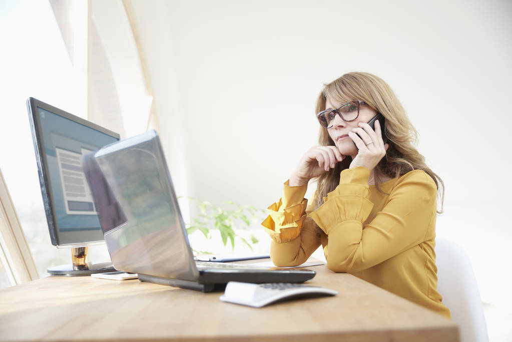 Bizneswoman średniej wieku konsultant za pomocą jej telefon komórkowy i rozmowy z klientem siedząc przy biurku przed laptop i zarządzanie jej działalności.  - Zdjęcie, obraz