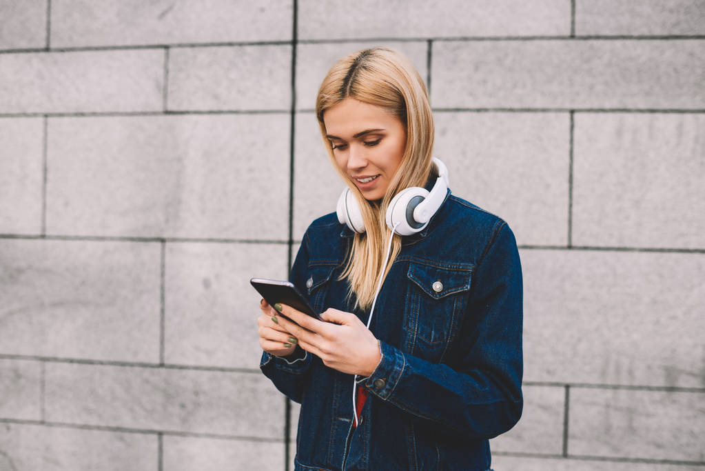 Blonde hipster girl in modern headphones on neck sharing media in social networks on smartphone connected to 4g internet.Female blogger chatting online on telephone on grey promotional background - Photo, Image