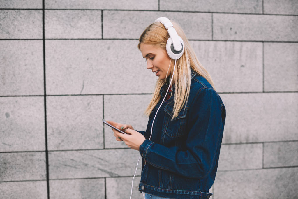 Zijaanzicht van de hipster meisje gekleed in denim kleding audio nummers in muziek toepassing op smartphone voor het luisteren in moderne hoofdtelefoon wandelen langs grijze promotionele achtergrond kiezen - Foto, afbeelding