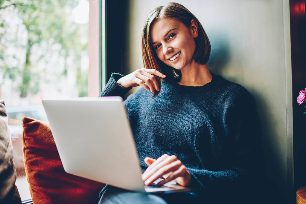 Portret hipster Ładna dziewczyna uśmiechając się aparatu podczas odpoczynku na parapecie w coworking z nowoczesny laptop komputer do poszukiwania informacji przydatnych dla rozwoju własną stronę internetową za pomocą darmowego wifi - Zdjęcie, obraz