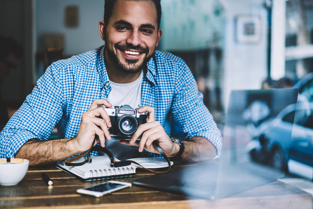 Πορτρέτο του χαρούμενα αρσενικό φωτογράφος που εργάζεται στο καφενείο εσωτερικών χρησιμοποιώντας σύγχρονο φορητό υπολογιστή και vintage φωτογραφική μηχανή για την κατασκευή εικόνες, ευτυχισμένο αρσενικό άτομο απολαμβάνοντας το χόμπι της φωτογραφίας αναπαύεται στο καφενείο - Φωτογραφία, εικόνα
