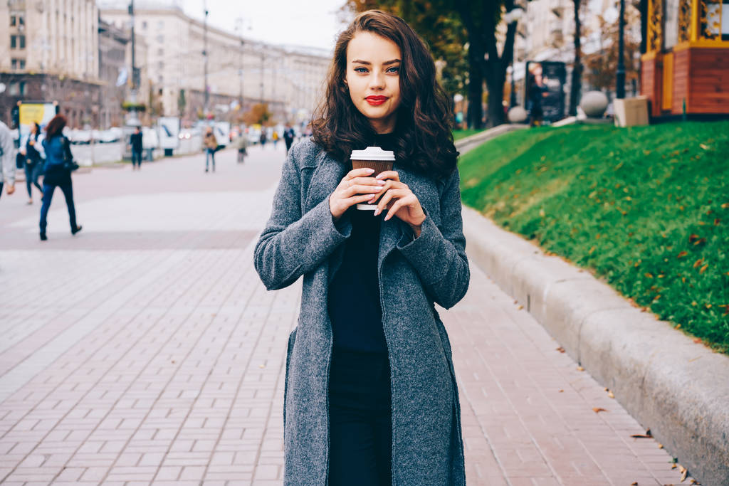 Portret van mooie jonge vrouw met rode lippen houden van lekkere koffie in de hand staan op straat in stedelijke omgevingen. Aantrekkelijke vrouw met warme drank camera buiten kijken. Promotionele achtergrond - Foto, afbeelding