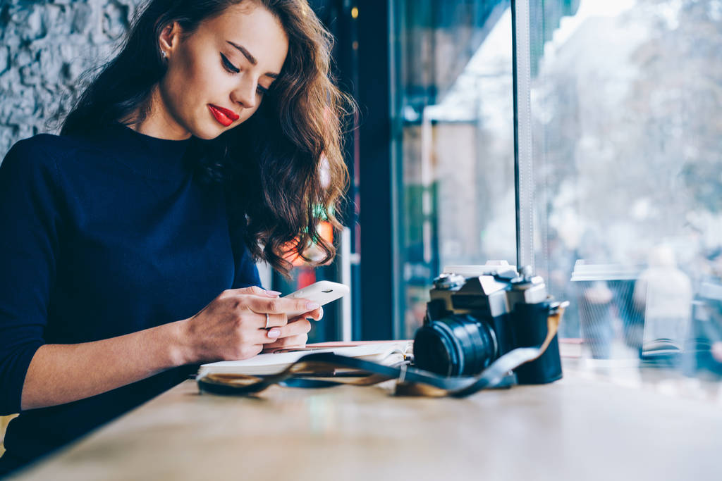 美しい若い女性の wifi ゾーンでコーヒー ショップ インテリア ビンテージ カメラでテーブルに座っている現代の携帯電話で、同僚とのオンライン チャットします。広告テキスト メッセージ用の領域をコピーします。 - 写真・画像