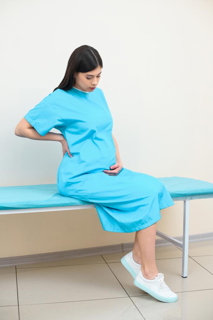 young pregnant woman sitting on bed at maternity hospital and holding her back - Photo, Image