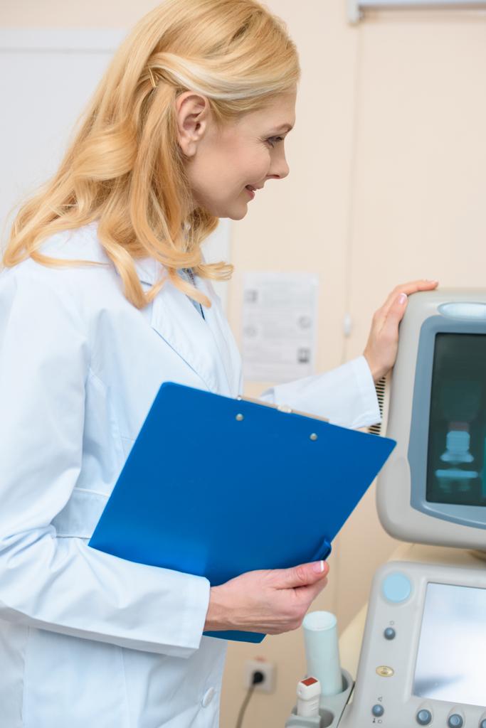 Lächelnder Arzt mit Klemmbrett, das Ultraschallscanner berührt - Foto, Bild