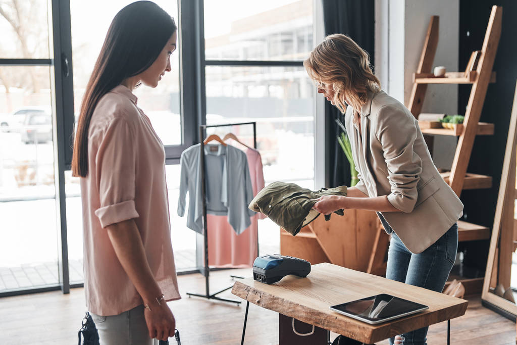 Verkäuferin faltet Hemd für Kundin, während sie in Modeboutique arbeitet - Foto, Bild