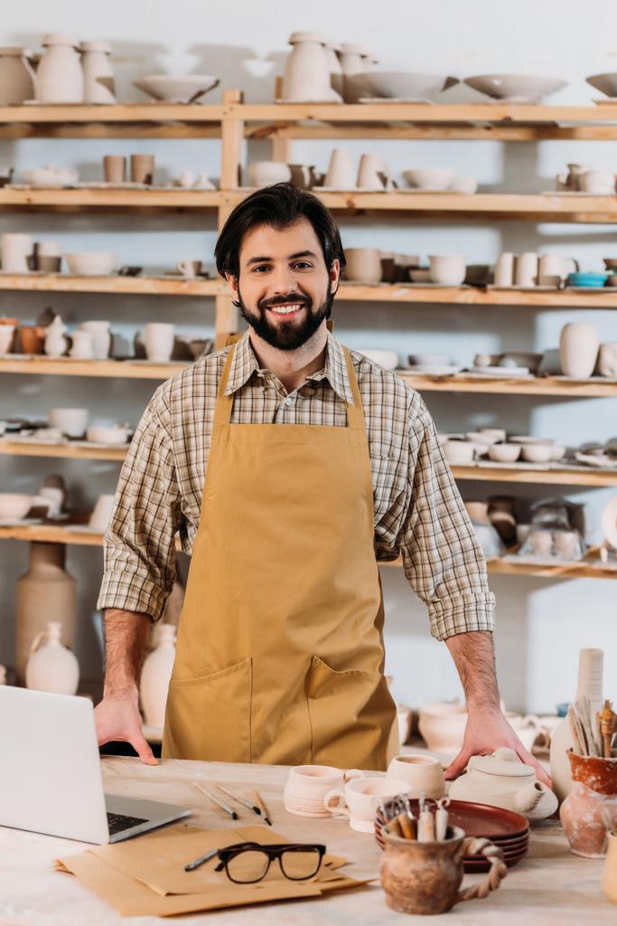 улыбающийся мужчина горшечник в фартуке, стоящий в мастерской с ноутбуком
 - Фото, изображение