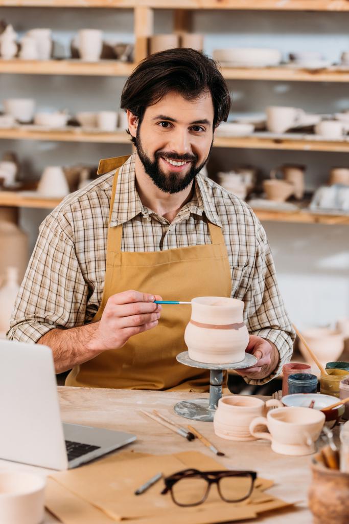 alfarero masculino feliz pintura cerámica vajilla en el taller con el ordenador portátil en la mesa
 - Foto, Imagen