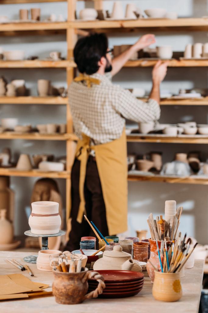セラミックの食器棚に立っている陶工のセレクティブ フォーカス ブラシし、塗料を前景上のテーブル - 写真・画像