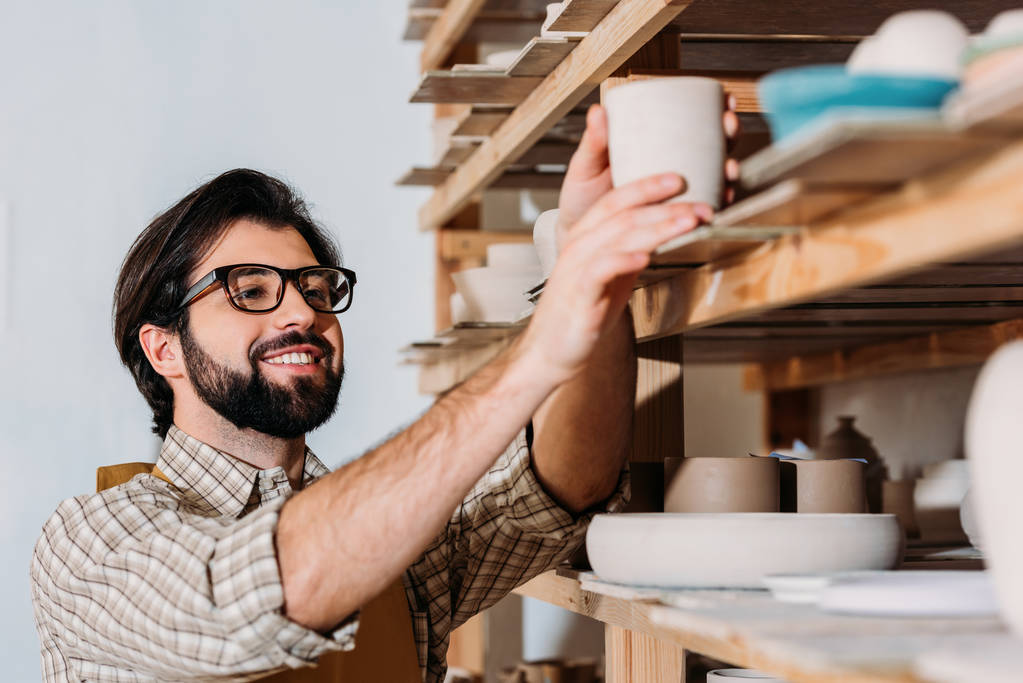 oleiro macho sorridente trabalhando com louça cerâmica em prateleiras na oficina
 - Foto, Imagem