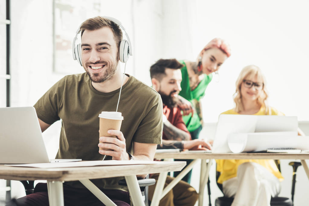 selectieve aandacht van glimlachende man in koptelefoon met koffie te gaan en creatieve collega's werken achter in kantoor - Foto, afbeelding