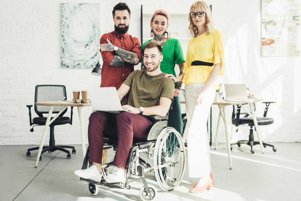 усміхнений інвалідний бізнесмен з ноутбуком і колегами, стоячи позаду в офісі
 - Фото, зображення