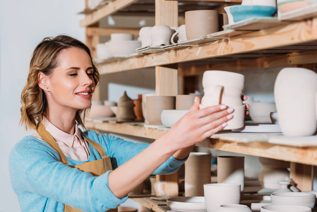 smiling potter with ceramic dishware on shelves in workshop - Photo, Image