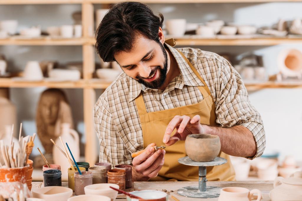 happy potter in apron decorating ceramic bowl in workshop - Photo, Image