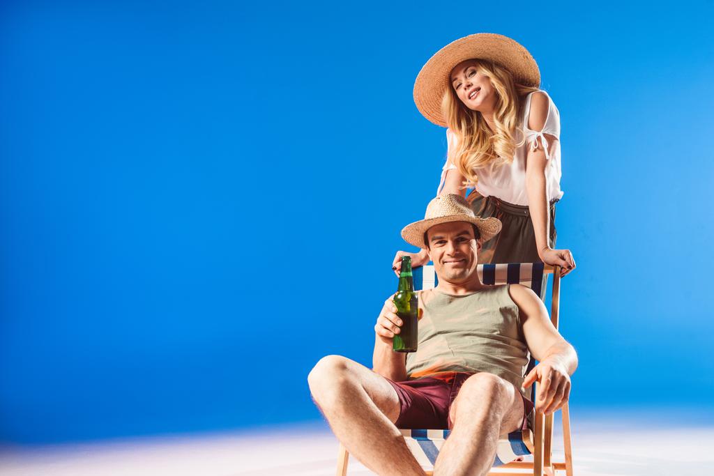 Blonde woman leaning on chair with man in straw hat holding beer on blue background - Photo, Image