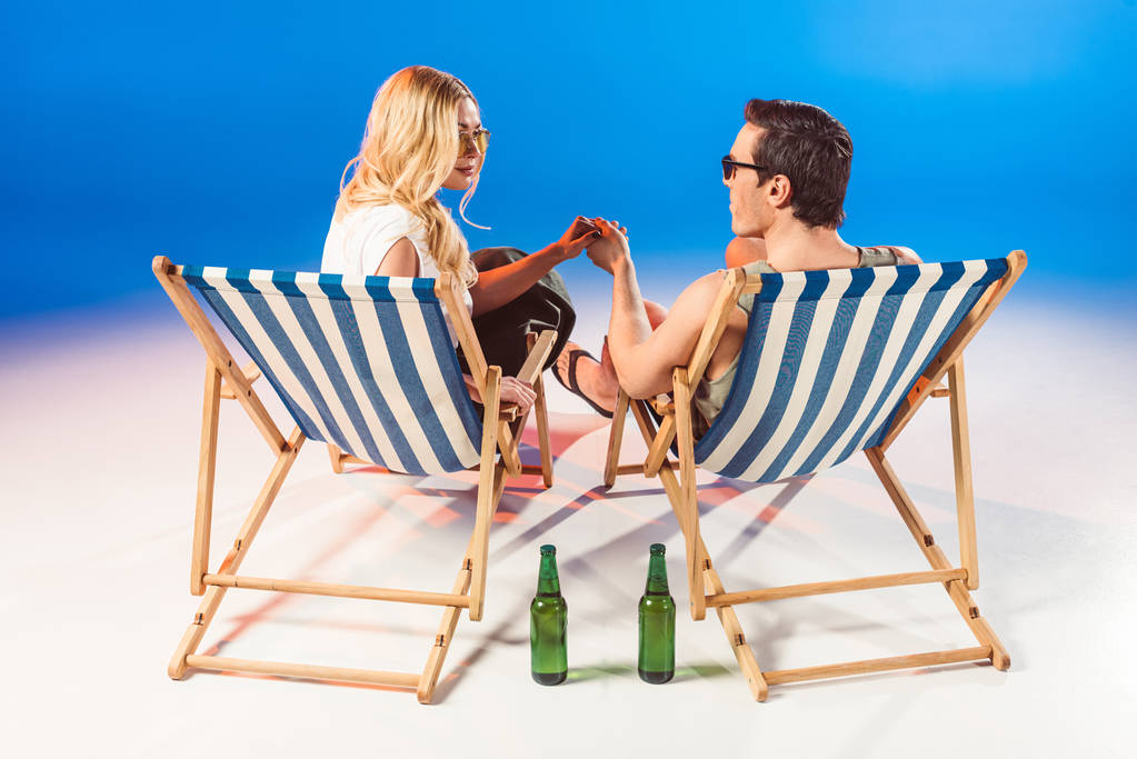 Jonge aantrekkelijke paar hand in hand in de ligstoelen door bierflessen op blauwe achtergrond - Foto, afbeelding