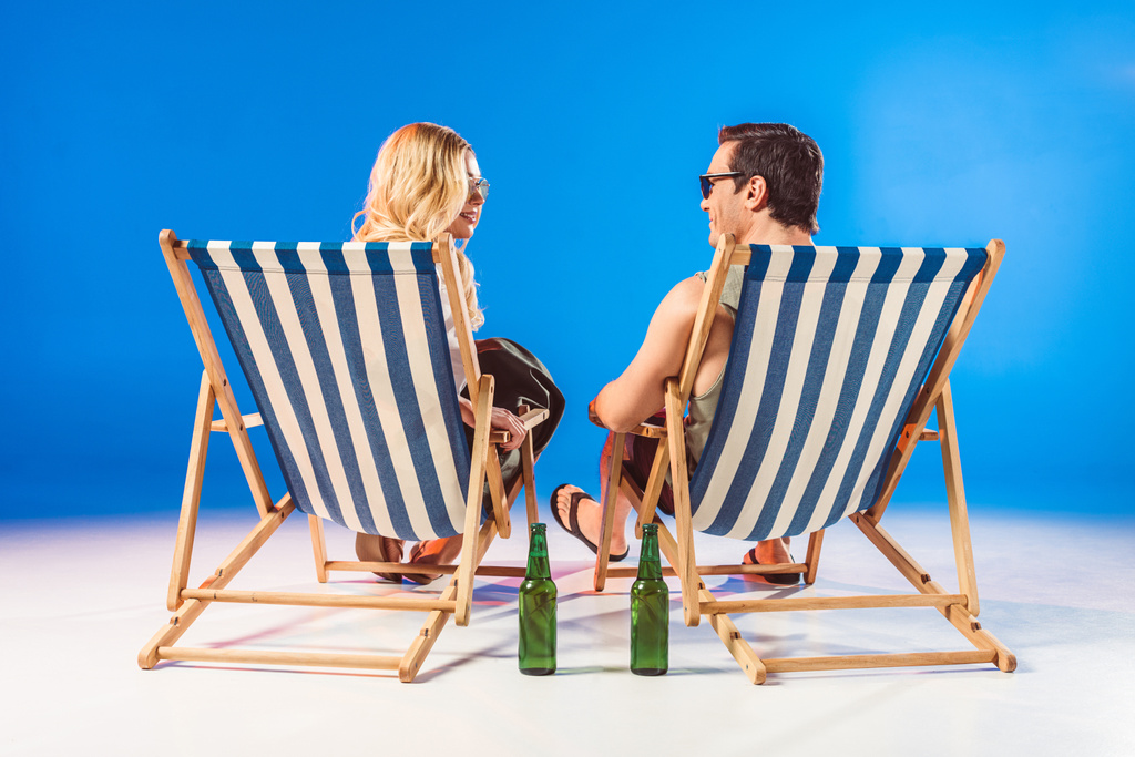 Smiling young couple relaxing in deck chairs by beer bottles on blue background - Photo, Image