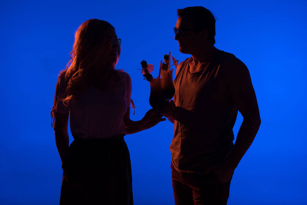 Hermosas siluetas de pareja tintineo con cócteles aislados en azul en la luz oscura
 - Foto, imagen