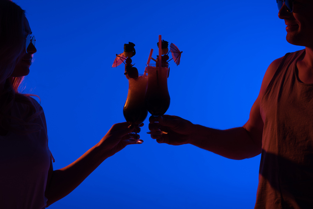 Homem e mulher brindam com coquetéis isolados em azul na luz escura
 - Foto, Imagem