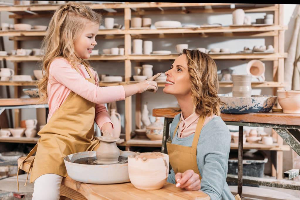 прелестная счастливая дочь трогает нос матери в мастерской по гончарному делу
 - Фото, изображение