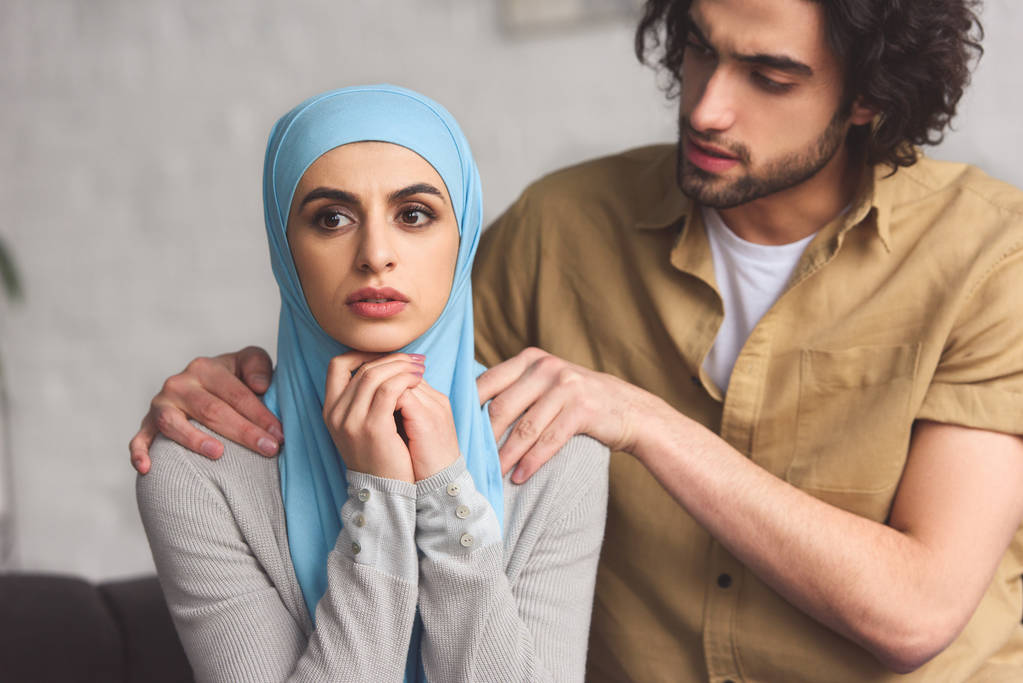Moslim vriendje knuffelen bezorgd vriendin in hijab in woonkamer - Foto, afbeelding