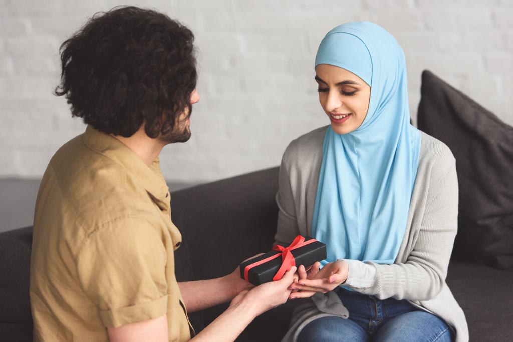 namorado muçulmano apresentando caixa de presente para namorada no hijab na sala de estar
 - Foto, Imagem