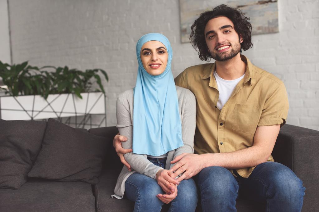sonriente pareja musulmana abrazando y mirando a la cámara en casa
 - Foto, Imagen