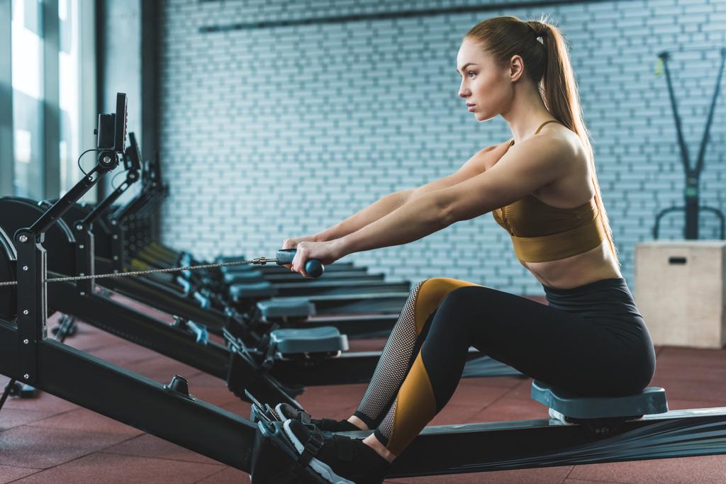 Side view of sportswoman doing exercise on rowing machine in sports center - Photo, Image
