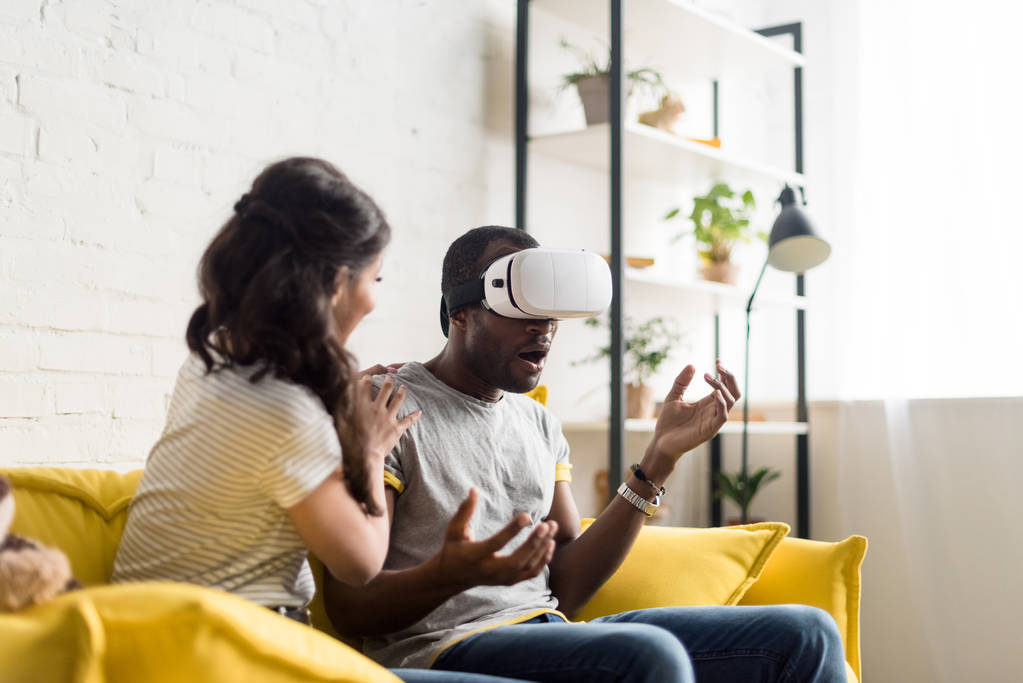 junge Afroamerikanerin sitzt neben schockiertem Freund, während er Virtual-Reality-Headset auf der Couch zu Hause trägt - Foto, Bild