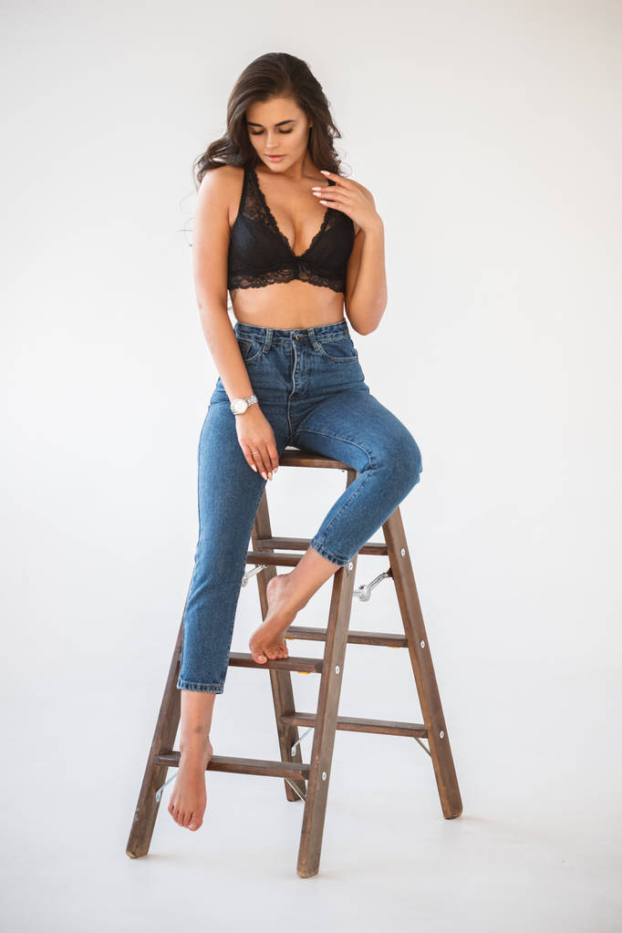 Studio ritratto di bella ragazza bruna riccia che indossa jeans blu e biancheria intima nera sessuale appoggiata su una scala di legno, sfondo bianco della parete
 - Foto, immagini