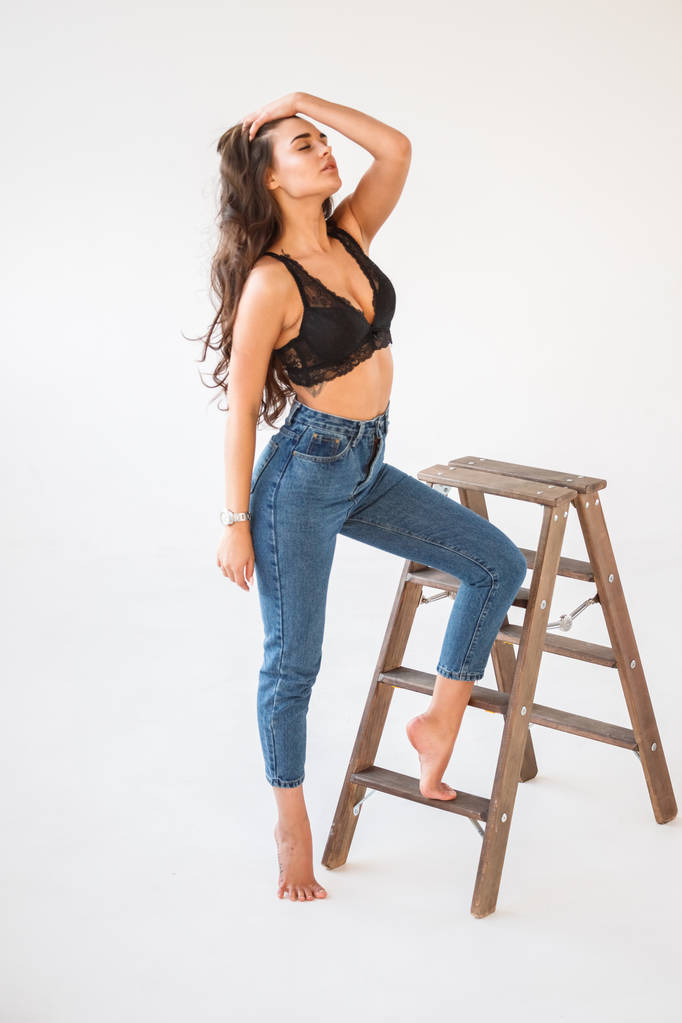 Studioporträt der schönen lockigen Brünetten, die blaue Jeans und sexuelle schwarze Unterwäsche trägt, angelehnt an eine Holztreppe, weißer Wandhintergrund - Foto, Bild