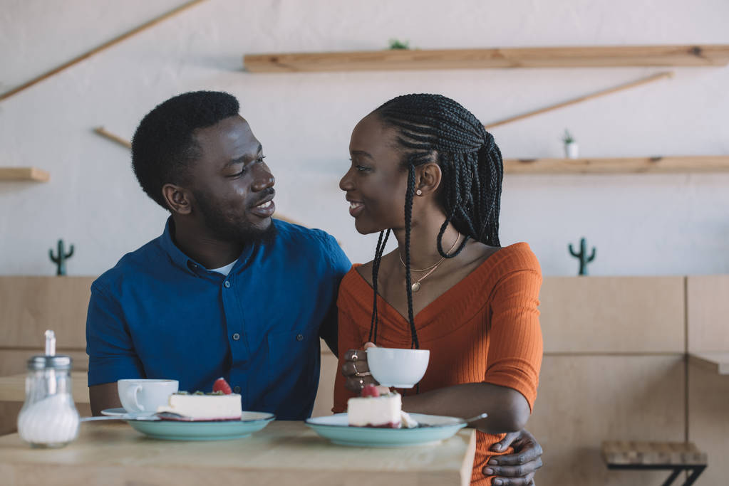 portrait of african american couple looking at each other while sitting at table with cups of coffee and desserts in cafe - Photo, Image