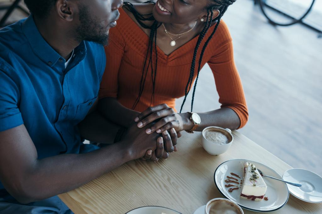 Teilansicht eines afrikanisch-amerikanischen Paares, das Händchen hält, während es bei einem romantischen Date im Café am Tisch sitzt - Foto, Bild