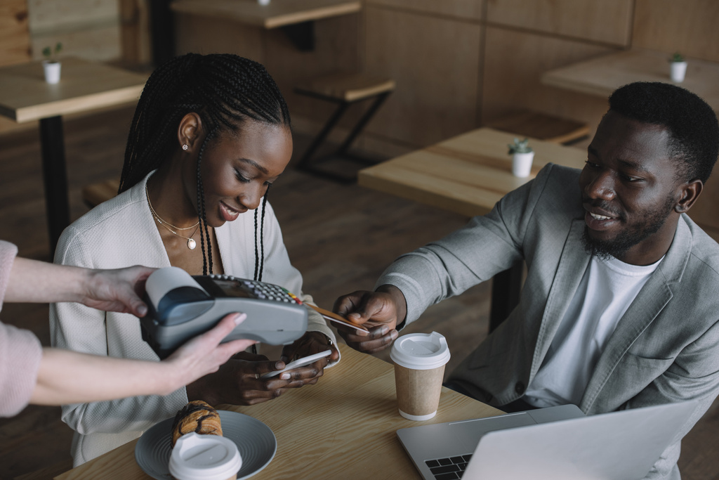 Afrikkalainen amerikkalainen mies maksaa tilauksen luottokortilla viettäessään aikaa ystävän kanssa kahvilassa
 - Valokuva, kuva