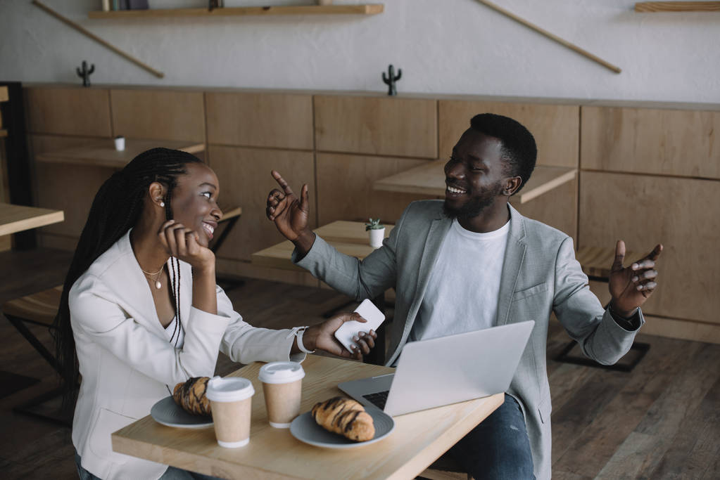 улыбающиеся африканские американские друзья за столом с ноутбуком в кафе
 - Фото, изображение