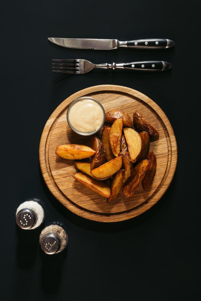 νόστιμες πατάτες φούρνου με σάλτσα σε ξύλινη σανίδα, μπαχαρικά και πιρούνι με μαχαίρι σε μαύρο  - Φωτογραφία, εικόνα