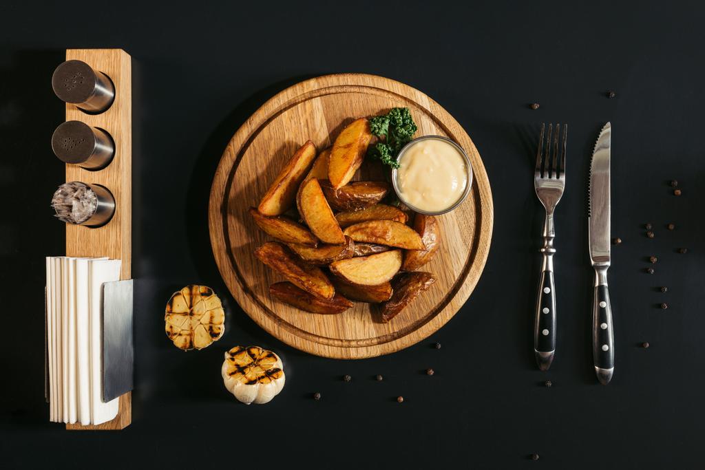 top view of delicious roasted potatoes with sauce on wooden board, grilled garlic and fork with knife on black - Photo, Image