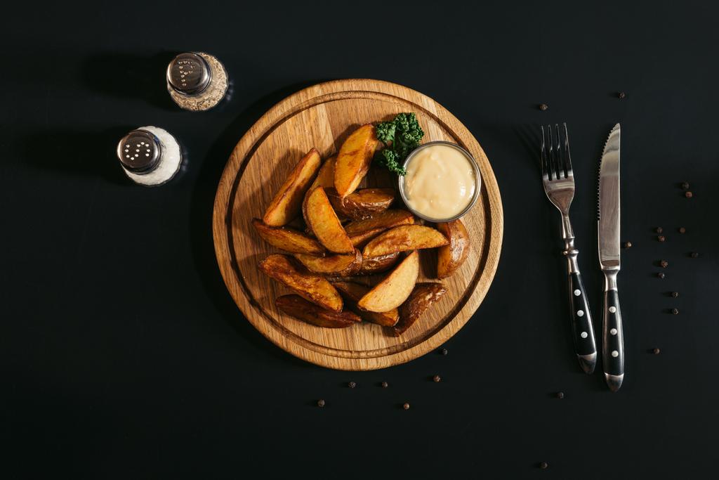 top view of baked potatoes with sauce on wooden board, fork with knife and spices on black - Photo, Image