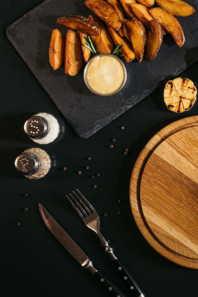 pohled shora na chutné pečené brambory s omáčkou, kořením, grilovaným česnekem a dřevěná deska s vidličkou a nožem na černém pozadí - Fotografie, Obrázek