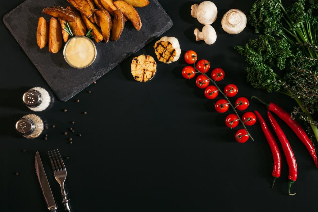 bovenaanzicht van lekkere gebakken aardappels met saus, kruiden en groenten op zwart - Foto, afbeelding