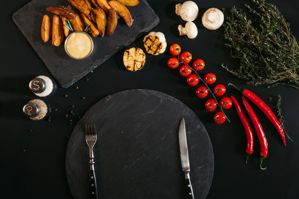 vue du dessus du panneau d'ardoise vide avec fourchette et couteau, pommes de terre cuites au four, épices et légumes sur noir
 - Photo, image