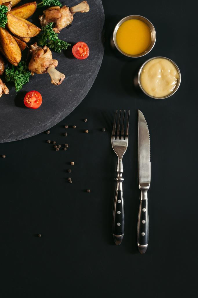 vue du dessus de la fourchette avec couteau et délicieuses pommes de terre rôties avec ailes de poulet et sauces sur noir
 - Photo, image