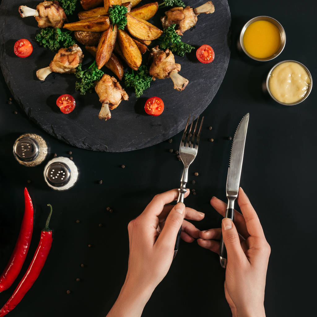 περικοπεί shot του προσώπου που κρατάει το πιρούνι και μαχαίρι ενώ τρώει νόστιμες ψητές πατάτες με κοτόπουλο και λαχανικά σε μαύρο - Φωτογραφία, εικόνα