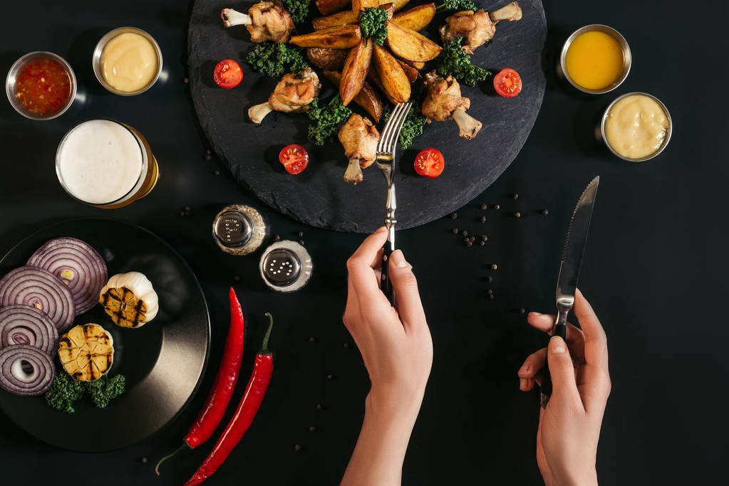 abgeschnittene Aufnahme einer Person, die Gabel und Messer in der Hand hält und leckere Ofenkartoffeln mit gebratenem Huhn und Gemüse auf schwarz isst - Foto, Bild