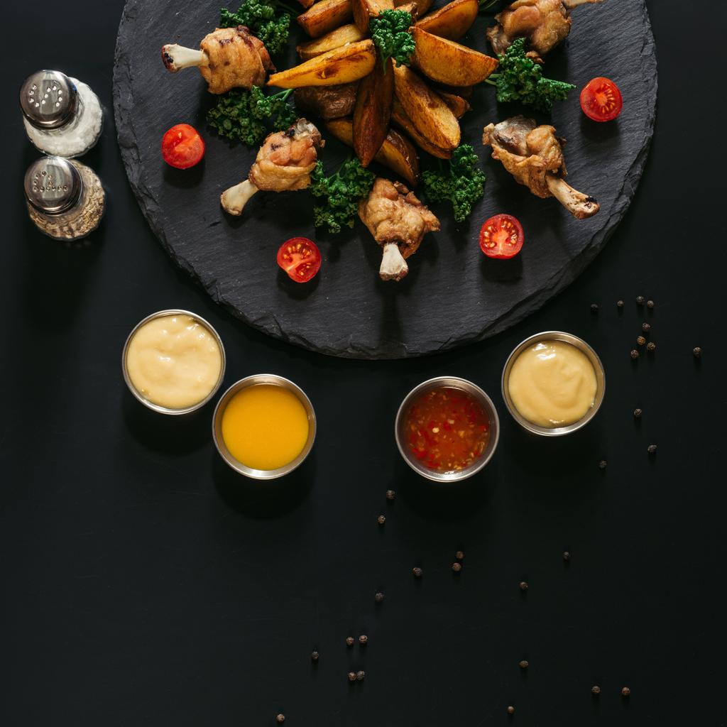 ylhäältä näkymä eri kastikkeet, mausteet ja herkullisia paistettuja perunoita paahdettuja kanansiipiä mustalla
  - Valokuva, kuva